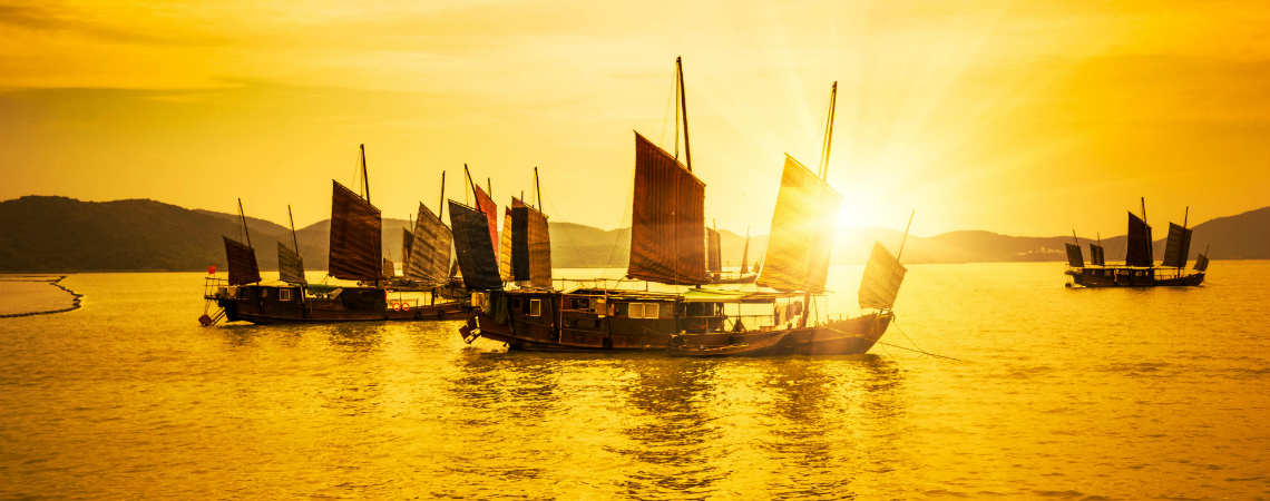 As the sun sets, Chinese junks are moored in a sheltered harbor.