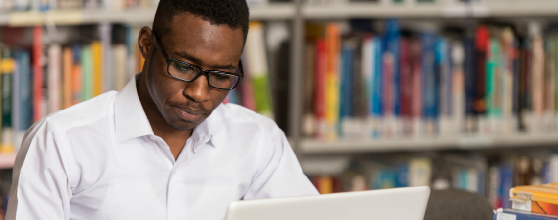 A young man wearing glasses works on a laptop in a library.
