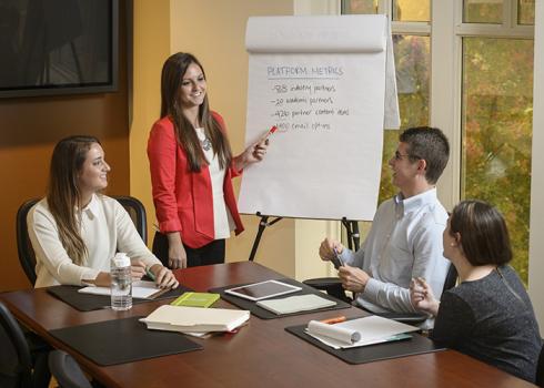 A group of young professionals gather around a conference table to listen to a flip-chart presentation.