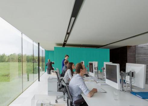 Employees sit at workstations along a long table in a sleek, modern office full of windows and natural light.
