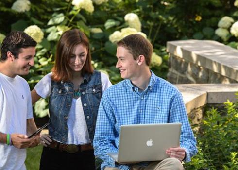 Three college students gather around a laptop while hydrangeas bloom in the background.
