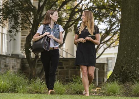 Two college women walk away from an academic building near a mature tree.