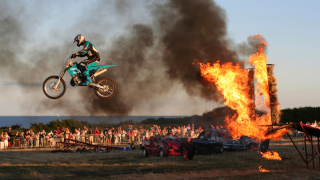 A crowd looks on as a motorcycle stunt driver soars through the air having jumped through a ring of fire.