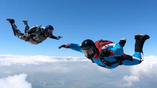 A pair of skydivers during freefall above the clouds. A river meanders in the distance.